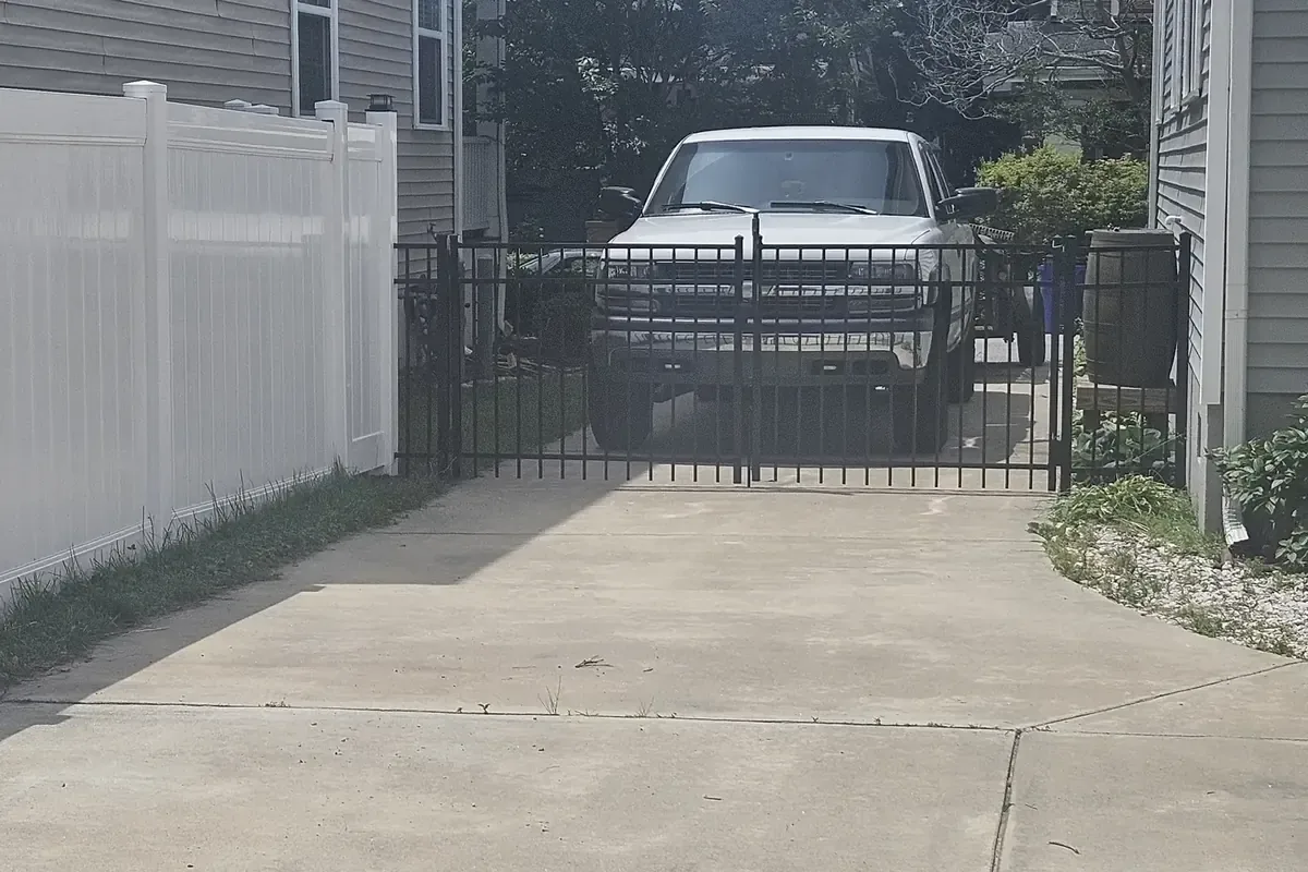 Black aluminum double drive gate across a concrete driveway, flat-top picket style, with a white vinyl privacy fence on the left, Chesapeake, Virginia